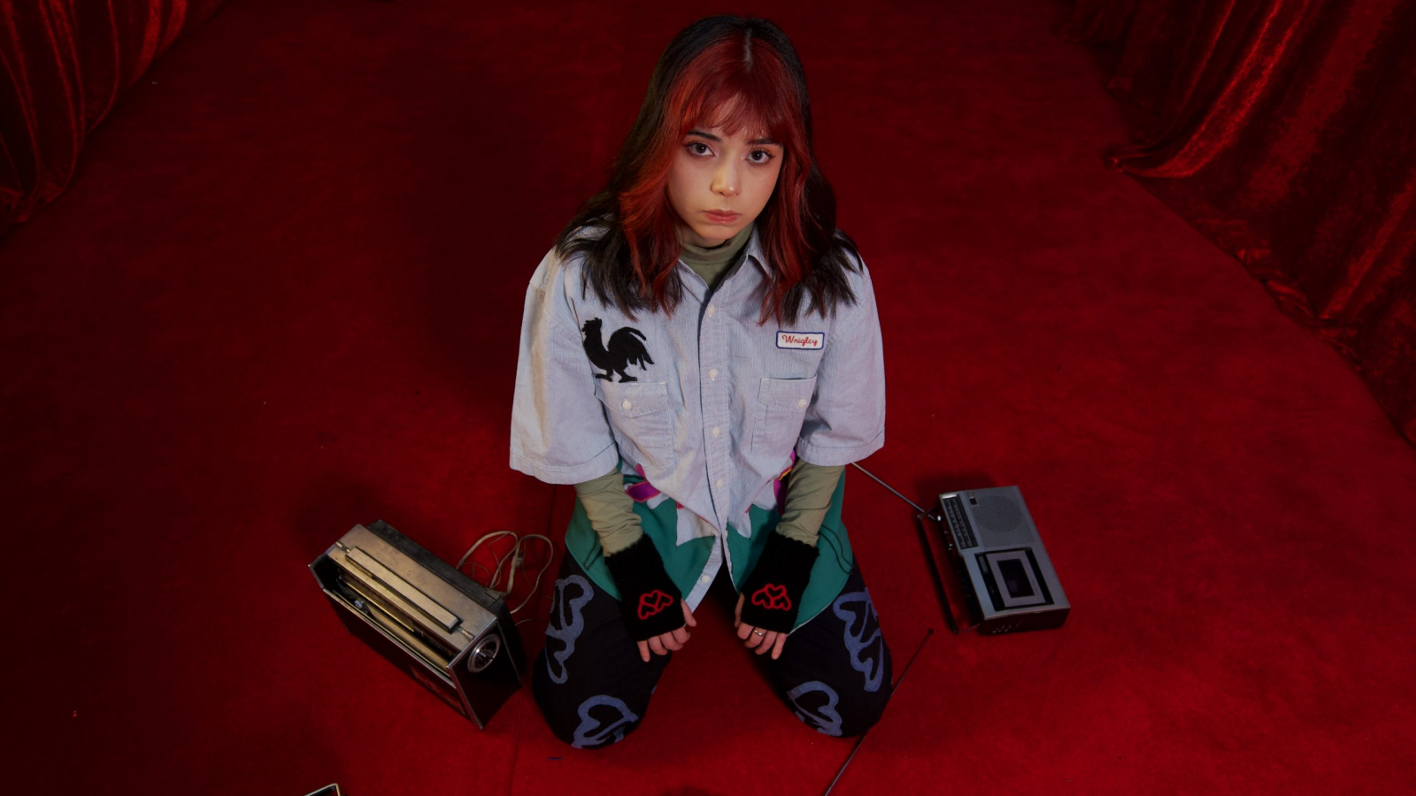 Bratty seated on a red set surrounded by cassette players, representing the dreamlike visual universe of her album and music video Radio