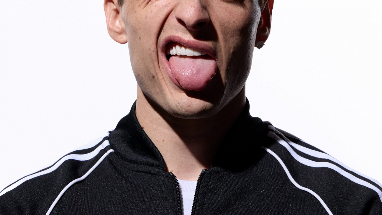 Peso Pluma con chamarra adidas Originals negra haciendo gesto irreverente mientras sostiene un objeto junto a su cabeza en retrato de estudio.