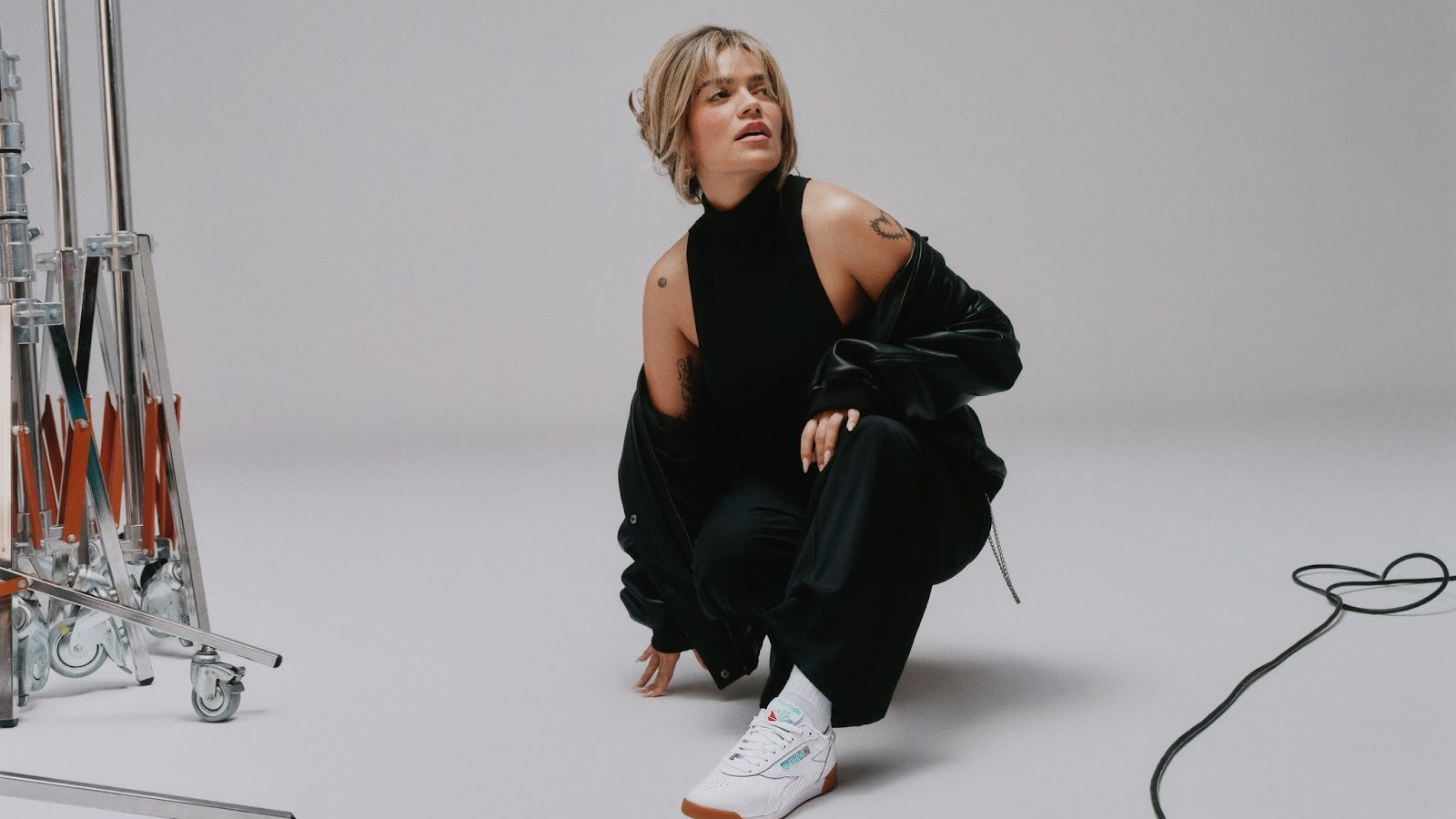 KAROL G posa en estudio con sneakers blancos Reebok Classic Workout Plus y outfit negro minimalista como parte de la campaña Born Classic Worn for Life 2026.