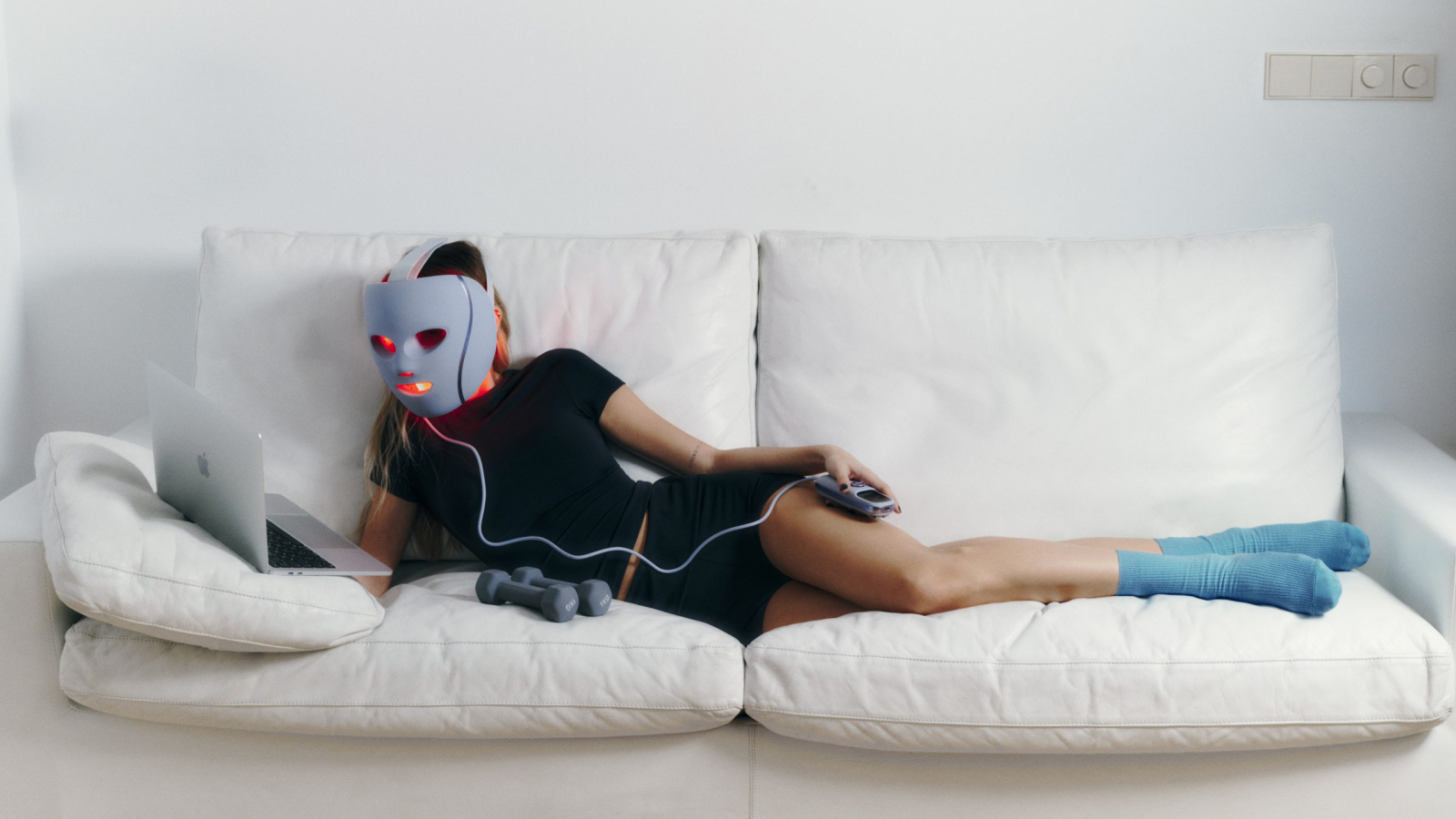 Modelo con look deportivo de Bershka Active recostada en un sofá blanco, usando máscara LED mientras sostiene un control y una laptop.