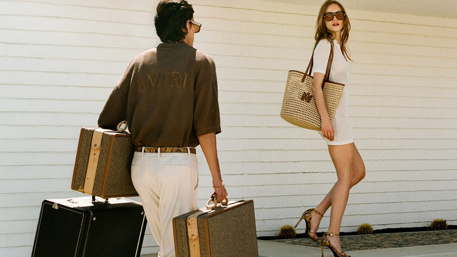 Hombre y mujer con looks de verano de AMIRI caminando con maletas en una casa de estilo mid-century bajo el sol de Palm Springs.