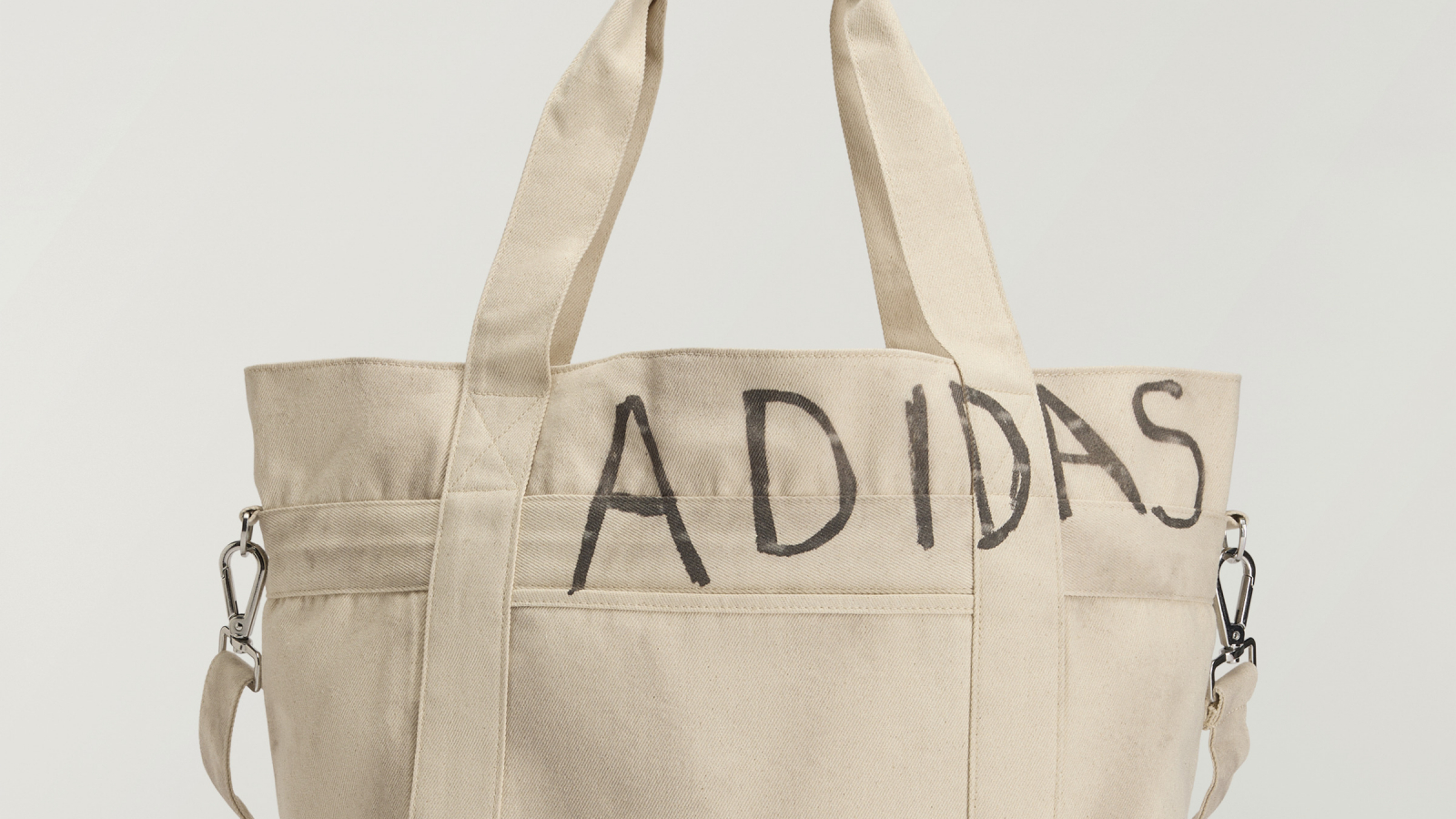 Tote bag de lona color beige de adidas Originals x Song for the Mute con gráfico manuscrito "ADIDAS" y diseño inspirado en la estética escolar y el desgaste cotidiano.