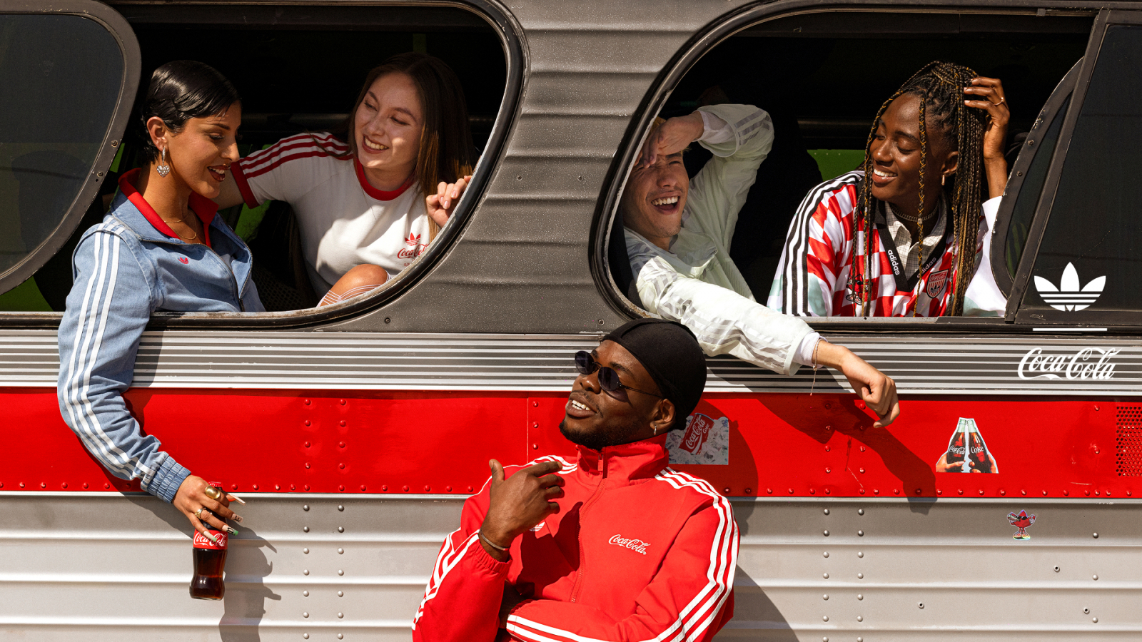 Grupo de jóvenes interactuando dentro y fuera de un autobús decorado con elementos de adidas Originals y Coca-Cola. Los participantes visten prendas de la colección colaborativa inspirada en la Copa Mundial de la FIFA 2026™, combinando estética deportiva retro, colores rojo, blanco y azul, y referencias visuales a ambas marcas.