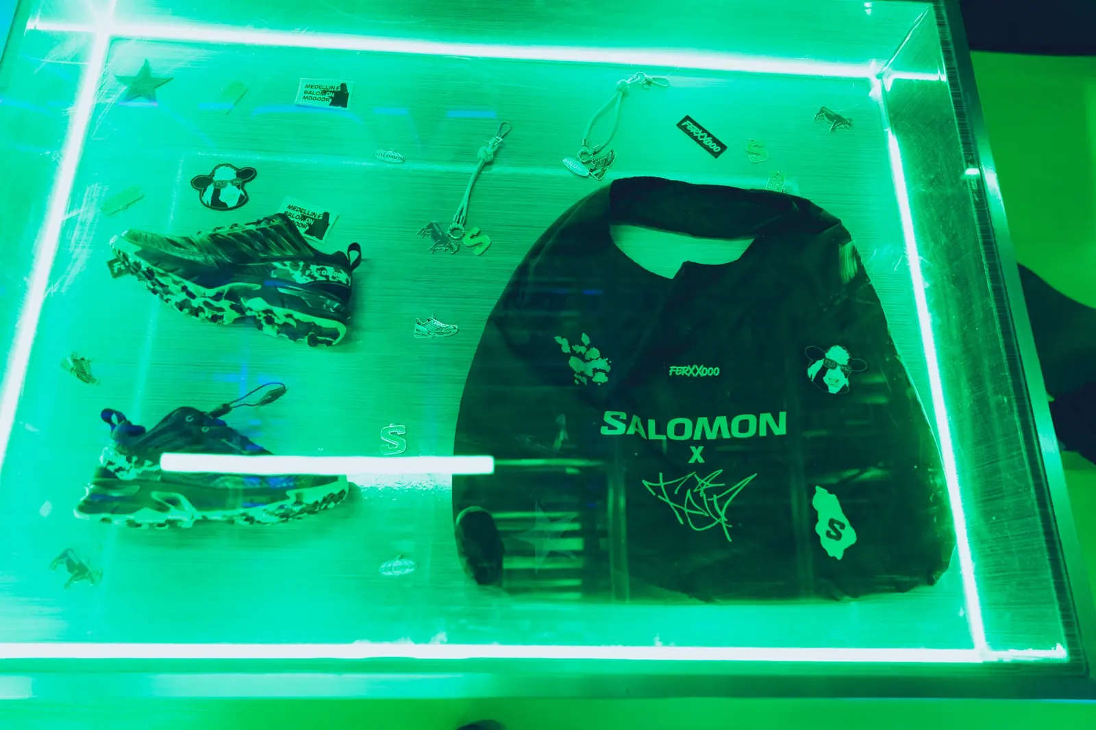 Vitrina iluminada en verde con los tenis The Green Star, accesorios y una camiseta con los logos de Salomon y Feid.