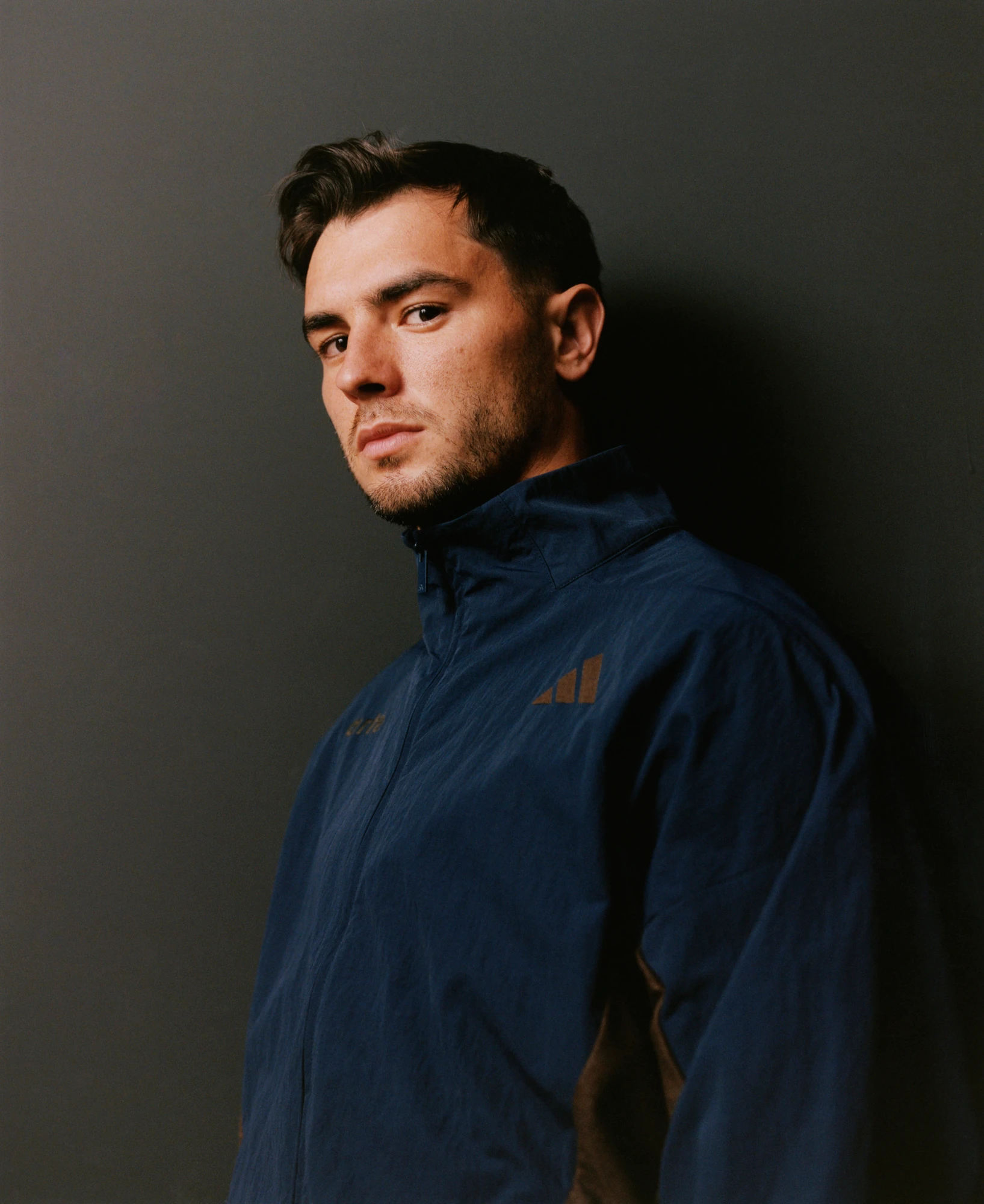 Retrato de campaña con una chamarra azul oscuro de cuello alto y logotipos de adidas x Arte Antwerp sobre un fondo gris.