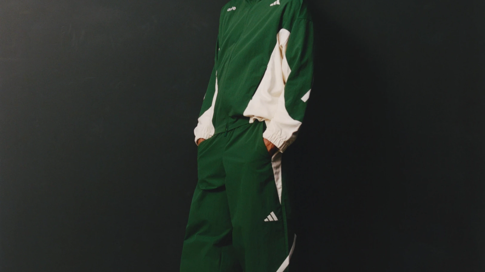 Look de adidas x Arte Antwerp con chamarra y pantalones verdes de paneles claros, acompañado de tenis blancos sobre un fondo gris oscuro.