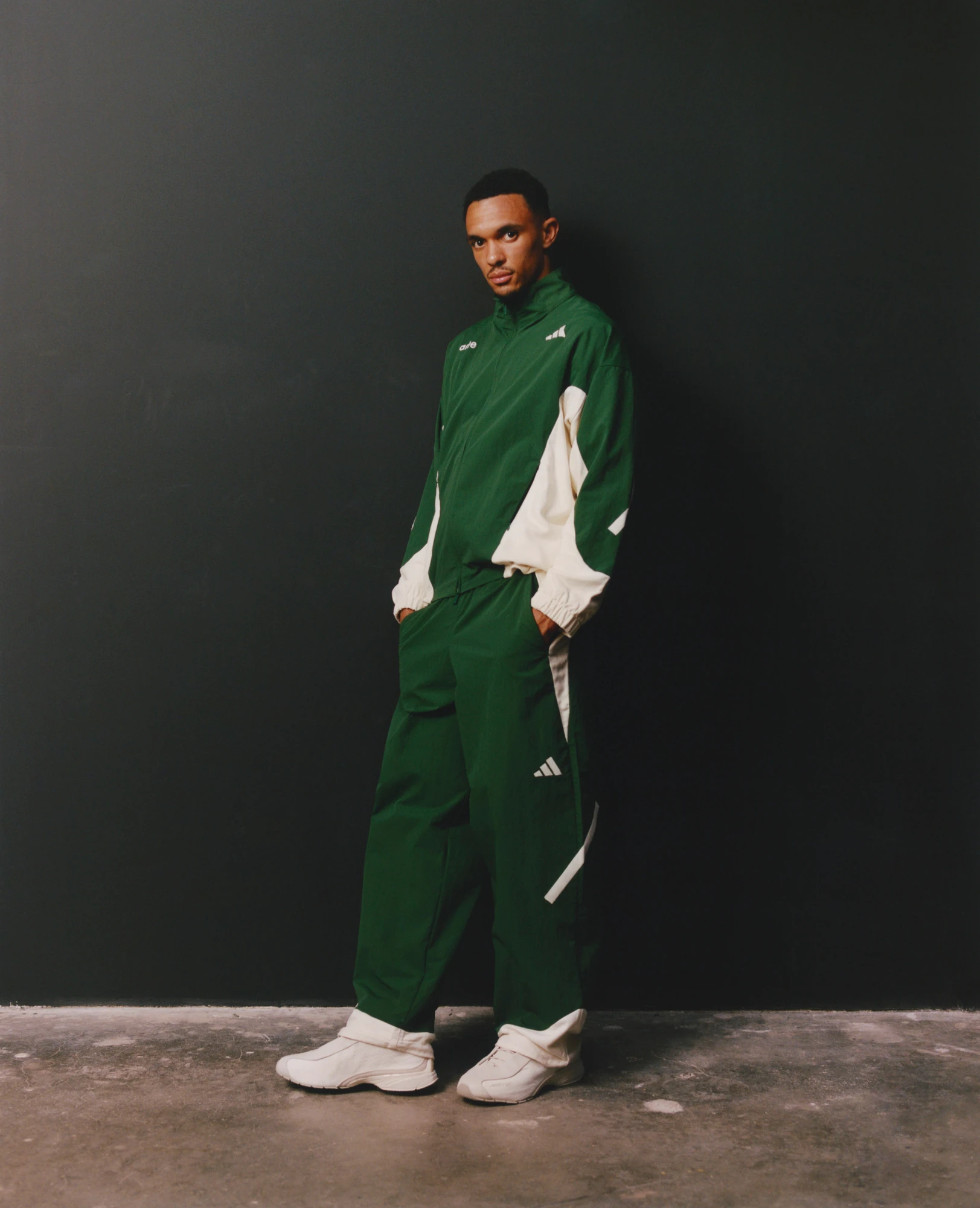 Look de adidas x Arte Antwerp con chamarra y pantalones verdes de paneles claros, acompañado de tenis blancos sobre un fondo gris oscuro.
