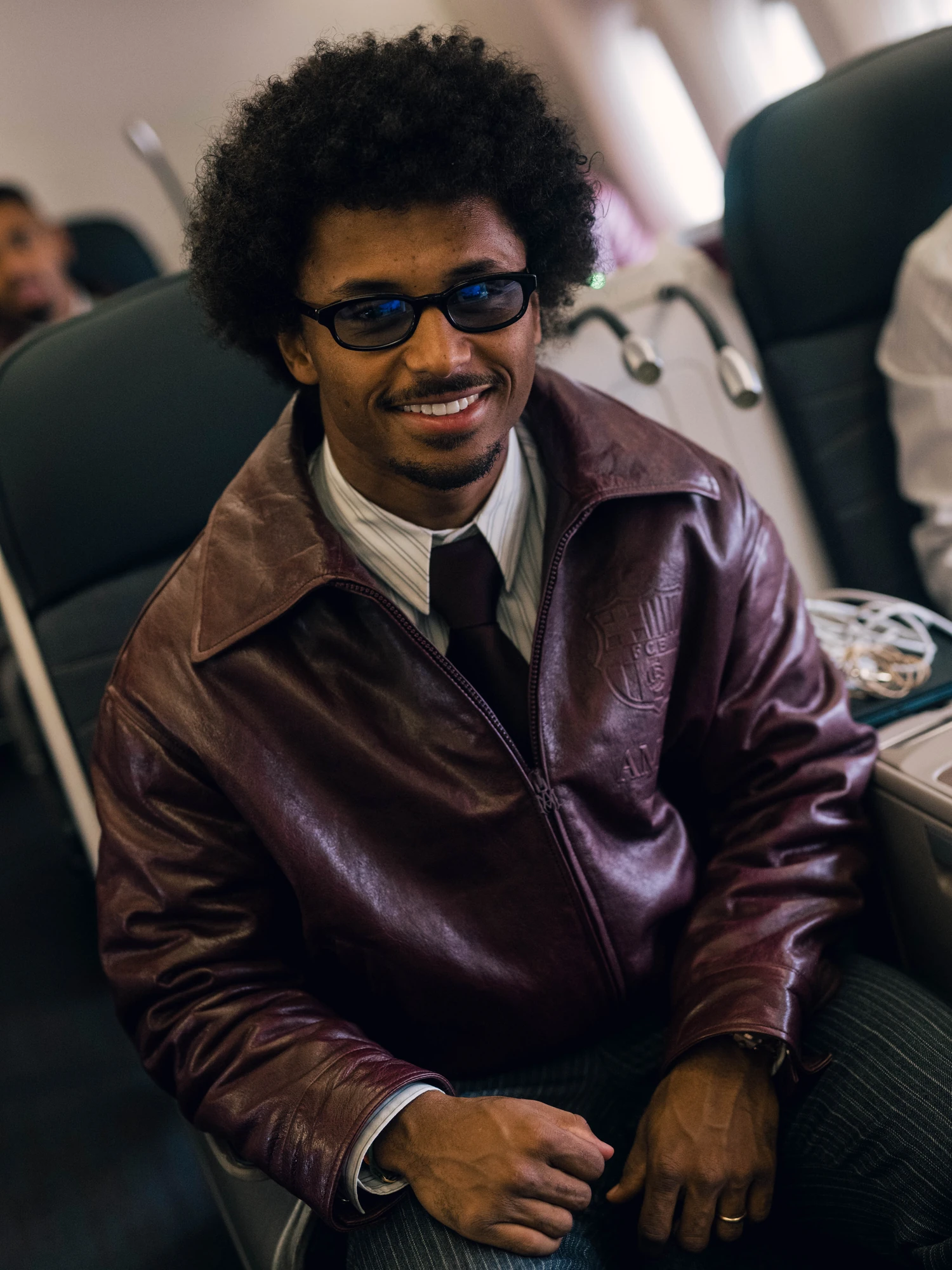 FC Barcelona player in a garnet leather AMIRI jacket over grey formalwear, seated inside an aircraft.