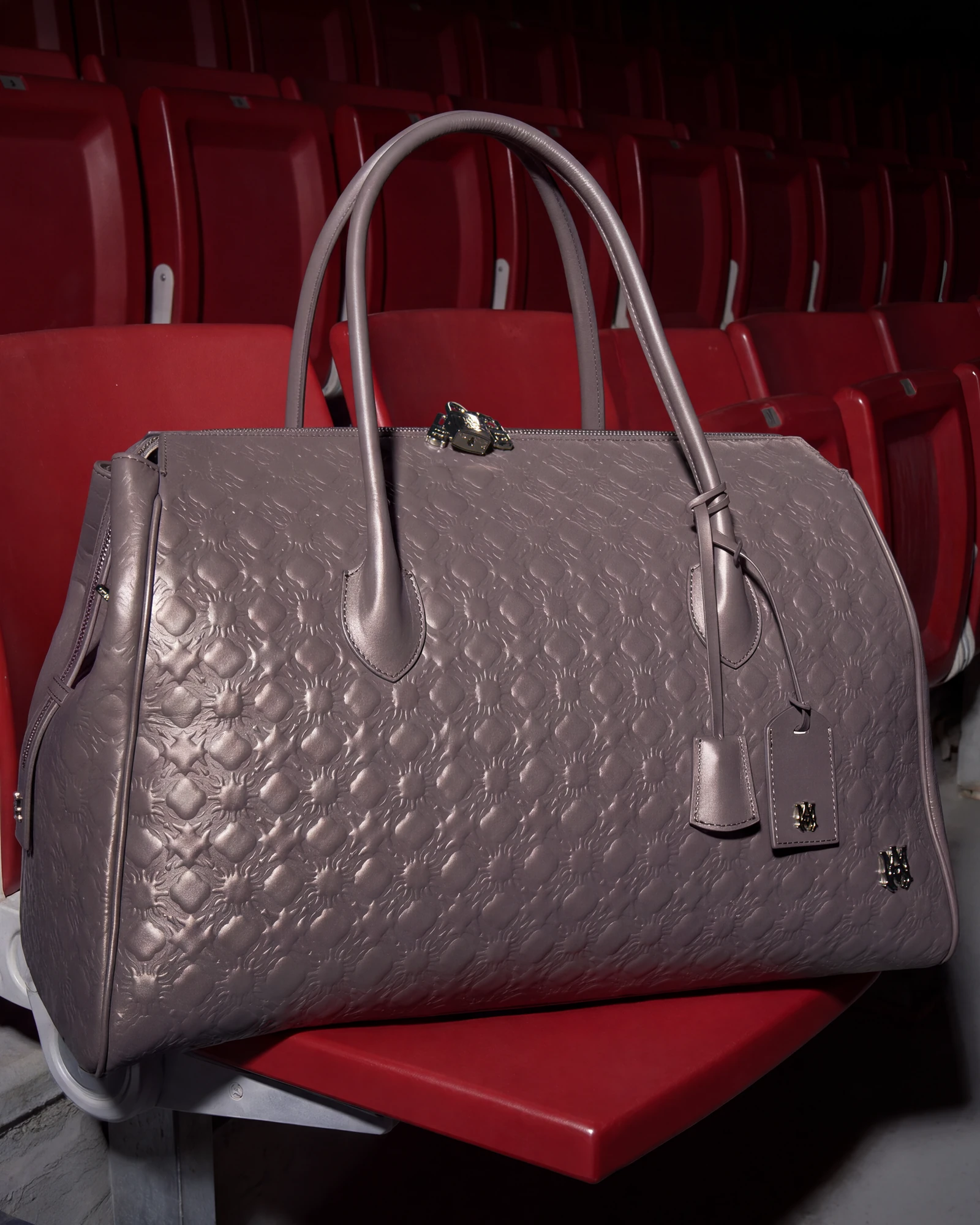 Grey AMIRI travel duffle with an embossed MA Quad Monogram resting on red stadium seats.