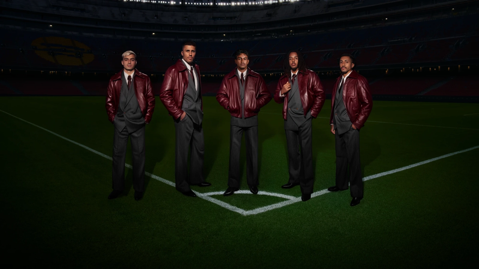 Five FC Barcelona players wearing AMIRI grey tailoring and garnet leather jackets on a stadium football pitch.