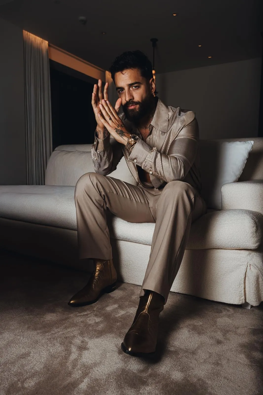 Maluma sentado en un sofá con camisa de rayas y pantalones en tonos neutros de AMIRI, mostrando las botas doradas Gold Ryder.