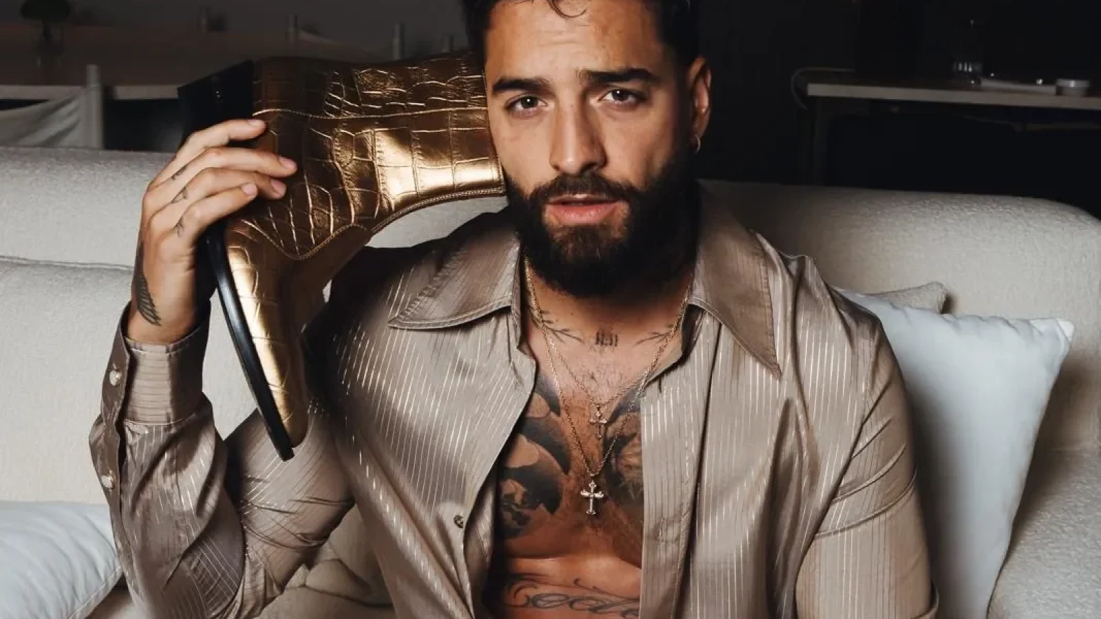 Maluma sentado en un sofá con una camisa de rayas AMIRI y pantalones en tonos neutros, sosteniendo una bota dorada Gold Ryder.