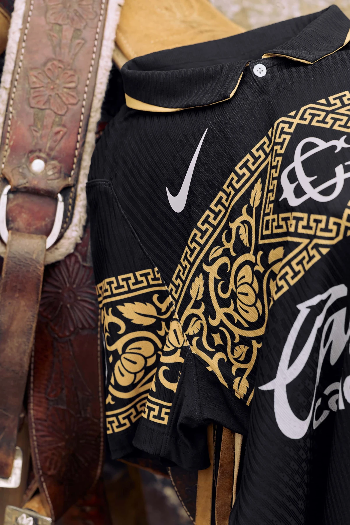Detalle del cuello, el escudo y el patrón dorado del jersey negro de Chivas junto a una silla de montar.