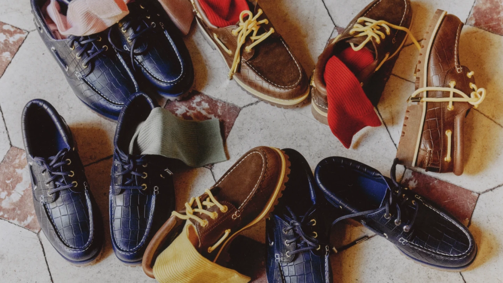 Náuticos Timberland x Jacquemus en azul marino y café, acompañados de calcetines de colores sobre un piso de baldosas.
