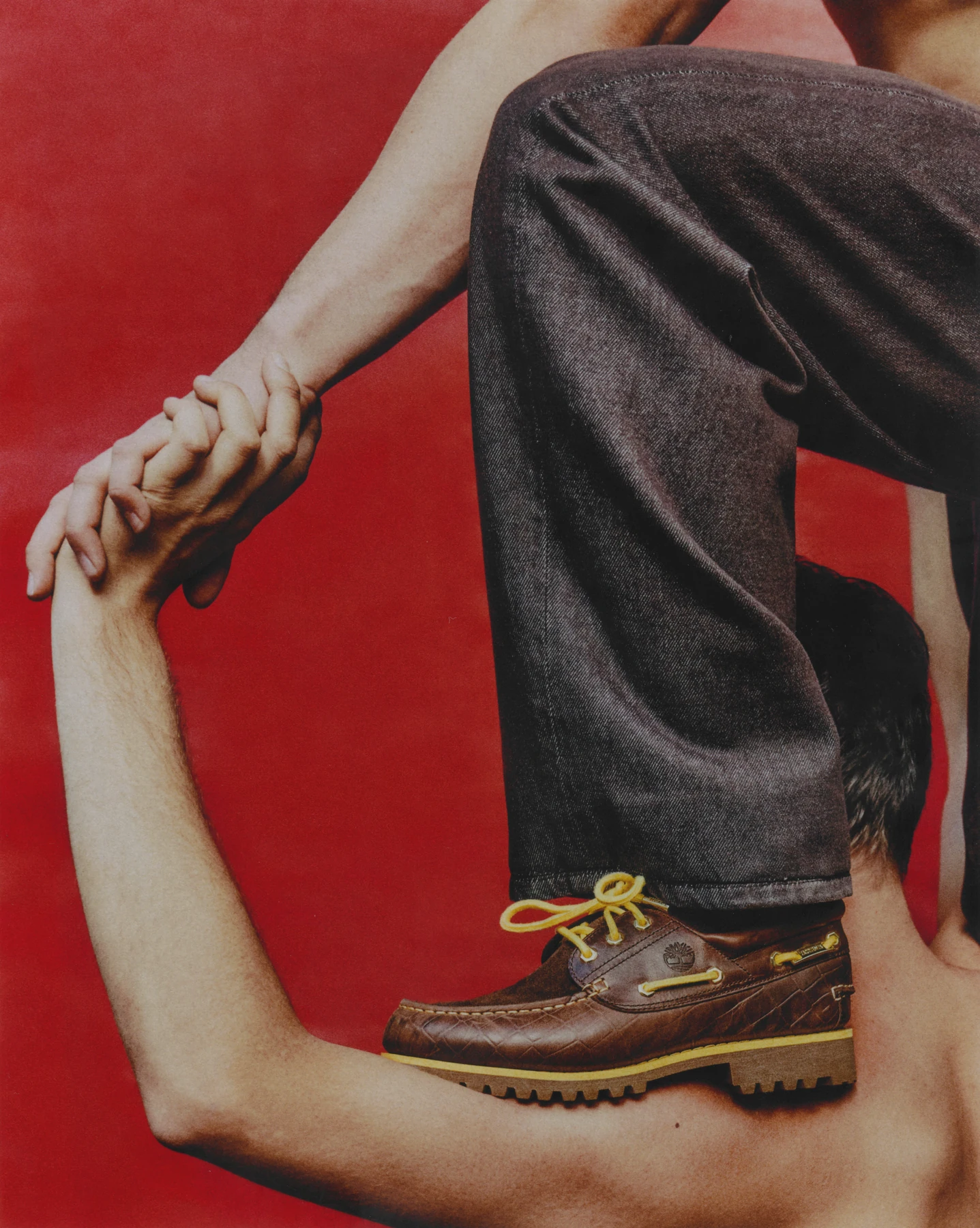 Composición de campaña con un náutico café de Timberland x Jacquemus, mezclilla y brazos sobre fondo rojo.