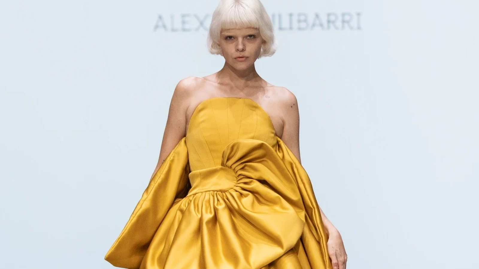 Modelo con vestido amarillo de hombros descubiertos, pliegues y volantes escalonados en la pasarela de Alexia Ulibarri.