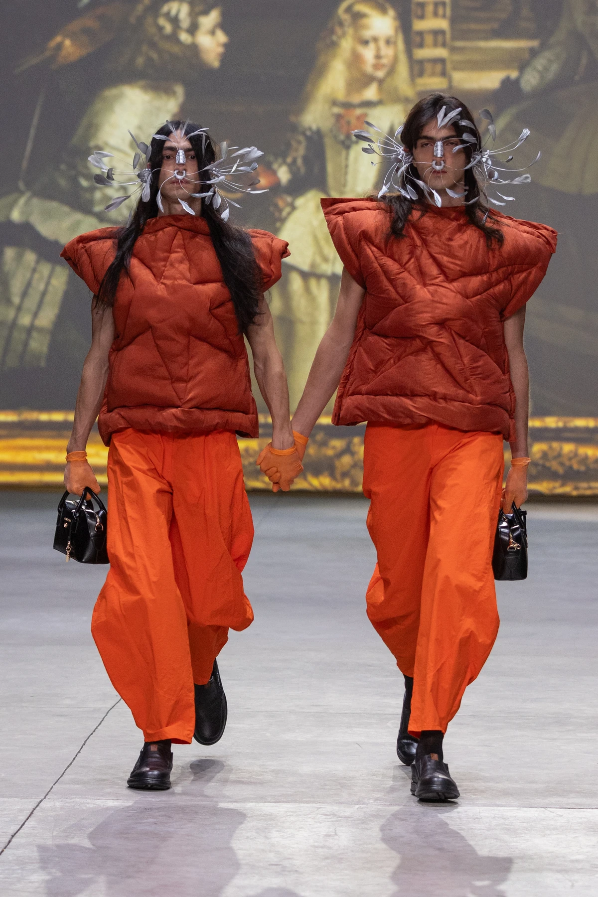 Dos modelos con prendas acolchadas rojizas, pantalones naranjas y adornos alrededor del rostro en el desfile de María Ponce.